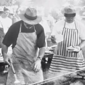 A vintage picture of two men wearing hats cooking food over a grill