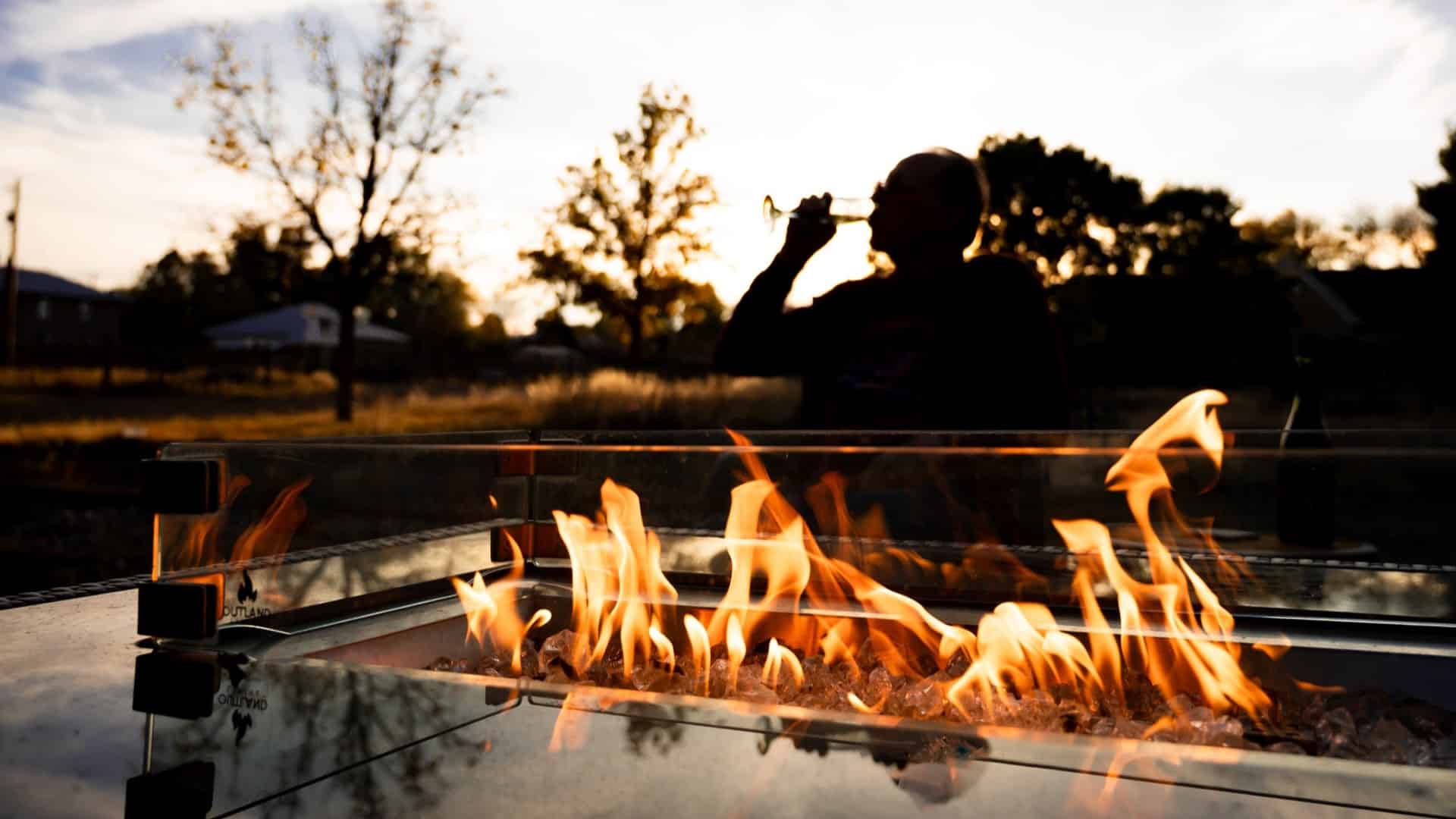 Picture of an outdoor firepit in low light or at sunset with a person behind it in silouette drinking from a champagne flute.