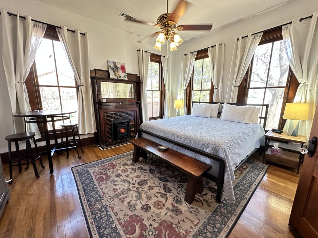 Elegant bedroom with king bed, fireplace, bistro table with two stools and large windows with white curtains