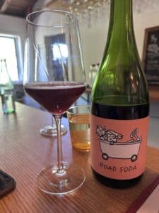 a botle of wine with a label that says Road Soda, a wine glass with red wine in it.