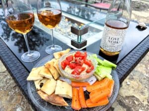 A bowl of dip covered with tomatoes surrounded by carrots, cucumber, and bread. The plate is on the edge of a fire pit and there are two glasses of rose wine and a wine bottle.