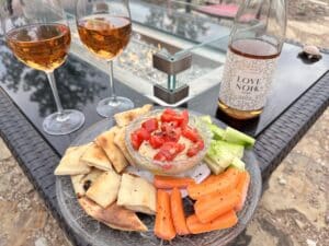 Two glasses of wine with rose wine, on a fire pit, with a plate of dip topped with tomatoes and surrounded by carrots, bread and cucumber slices. There's a bottle of Love Noir wine as well.