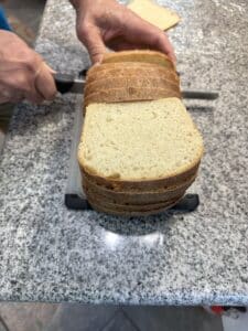 A loaf of bread being sliced. A stack of already sliced pieces are in the front of the photo with a hand holding a knife cutting the rest of the loaf behind.