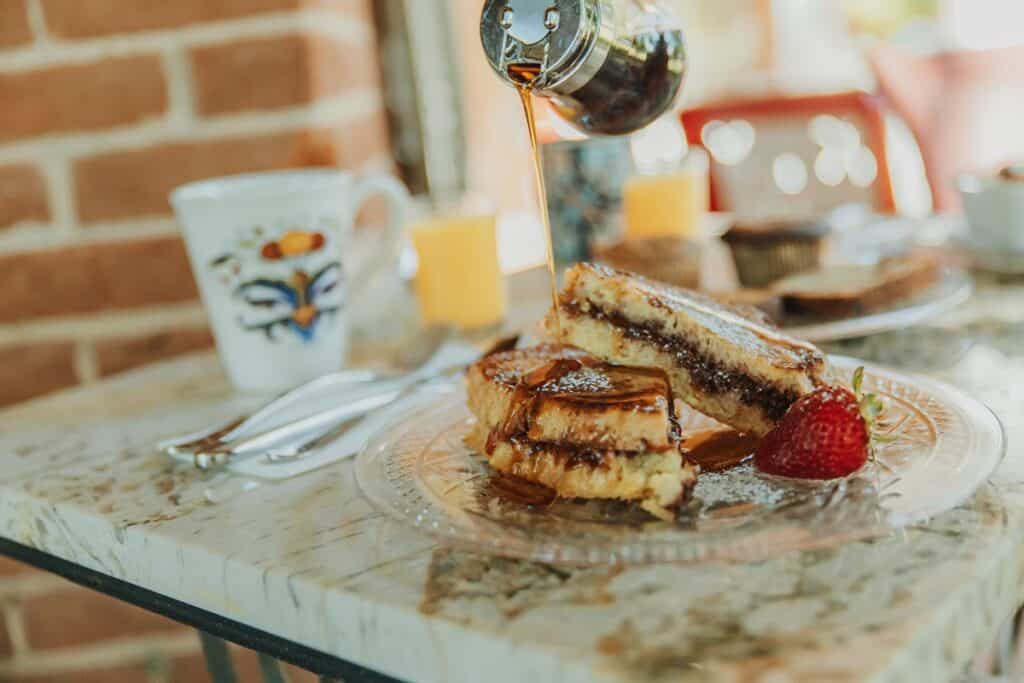 an outside table set with coffee, juice and a plate of french toast with someone pouring syrup over the plate.