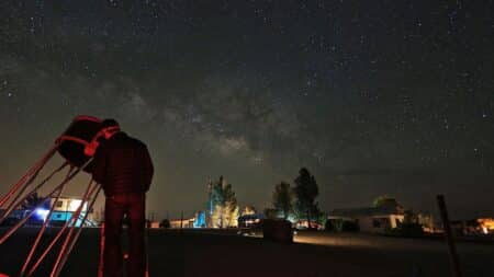 a person standing by a large telescope, looking into it. a dark sky is in the background with lots of stars.